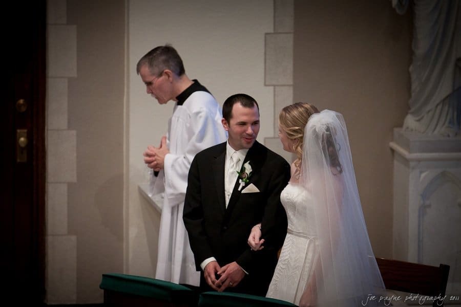 A bride and groom stand together in a church, smiling at each other. The groom is in a black suit, and the bride wears a white dress with a veil. A clergyman stands nearby, clasping his hands in prayer.