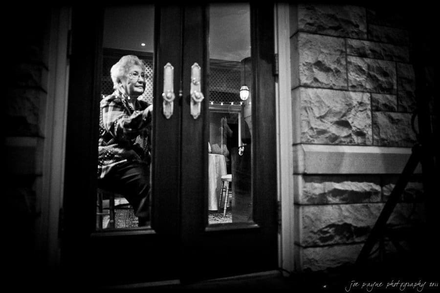 sacred heart cathedral wedding raleigh | monica and mark An elderly person sitting in a dimly lit room, viewed through glass double doors. The interior is softly illuminated, with some tables and chairs visible in the background. The person wears a patterned shirt and appears contemplative.