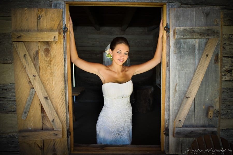 A woman in a white wedding dress stands in a wooden window frame, holding the shutters open. She has flowers in her hair and is smiling, surrounded by rustic wood textures.