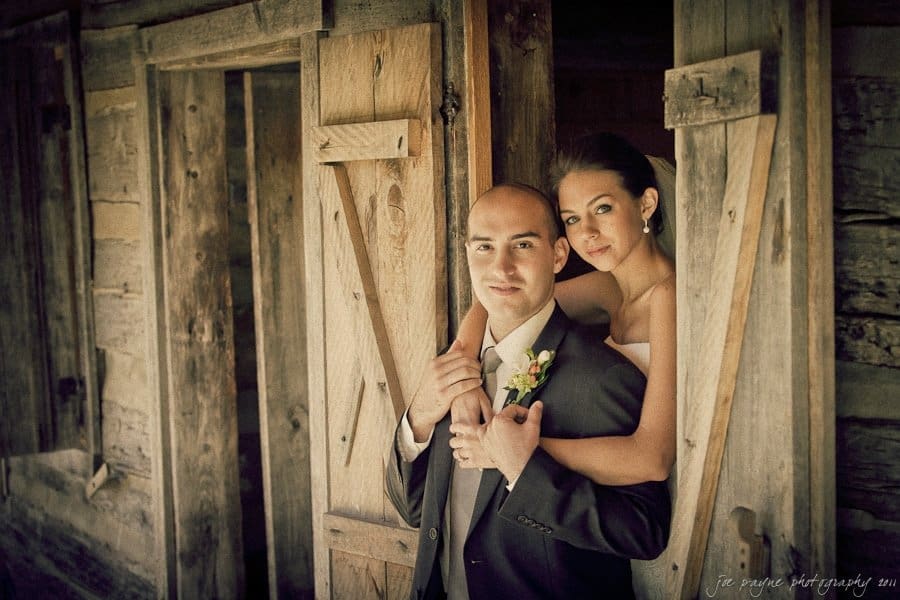 A couple stands in a rustic wooden doorway. The man in a suit holds the womans hand over his shoulder. The woman, in a white dress, leans close with a smile. The background features weathered wooden walls.