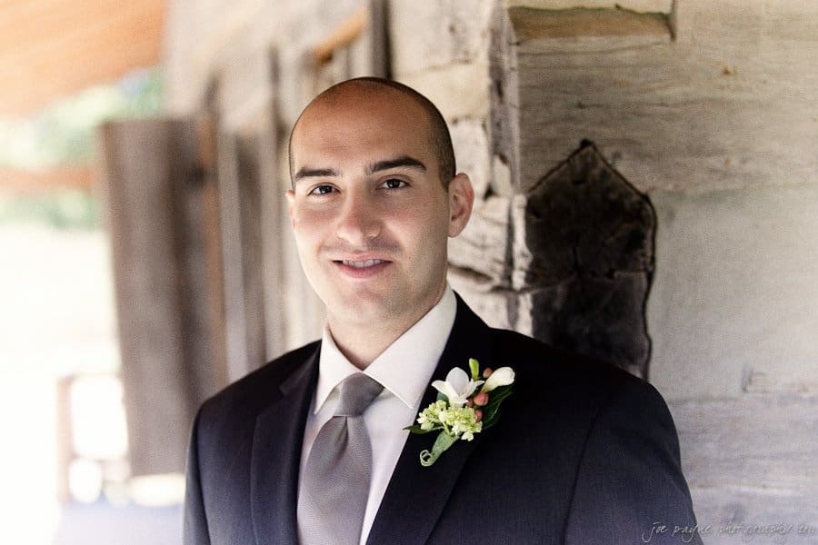 A man in a suit and tie, wearing a boutonniere with white flowers and greenery, stands outdoors next to a log building. He is looking at the camera with a slight smile.