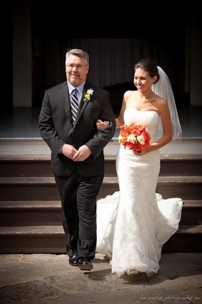 A bride in a white wedding dress holds a bouquet of orange and white flowers. She is smiling while walking arm-in-arm with a man in a black suit and tie. They are descending a set of steps, presumably at a wedding venue.