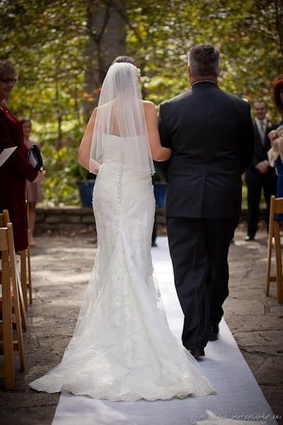 A bride in a white dress and veil walks down an outdoor aisle on a white runner, accompanied by a man in a dark suit. Guests are seated on wooden chairs, and trees are visible in the background.
