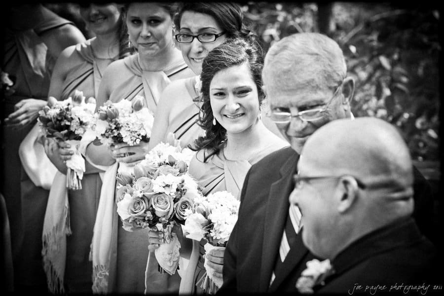 A black-and-white photo of a wedding ceremony. A bride smiles while standing beside bridesmaids holding floral bouquets. Two men in suits converse nearby. The mood is joyful and celebratory.