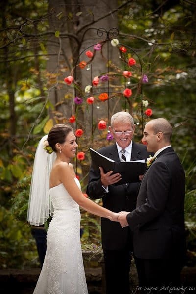 A bride and groom stand outdoors holding hands during their wedding ceremony. A smiling officiant reads from a book. The setting is decorated with colorful flowers and green foliage. The bride wears a white dress and veil, and the groom is in a black suit.