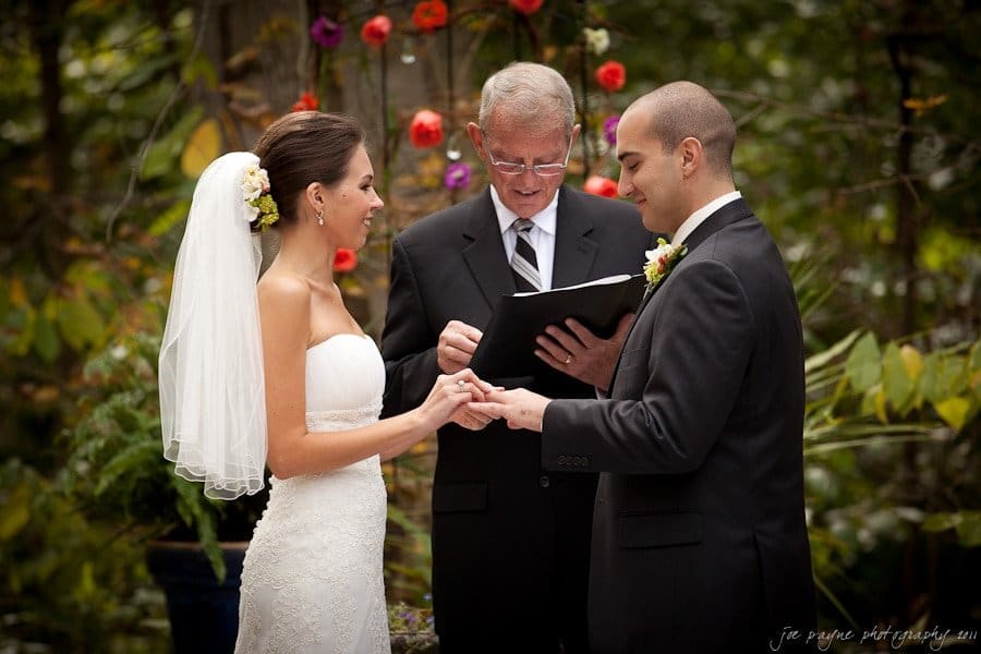 A bride and groom exchange rings during an outdoor wedding ceremony. The bride wears a white dress and veil, adorned with a floral hairpiece. The groom is in a dark suit. An officiant stands between them, holding a book. Colorful flowers are in the background.