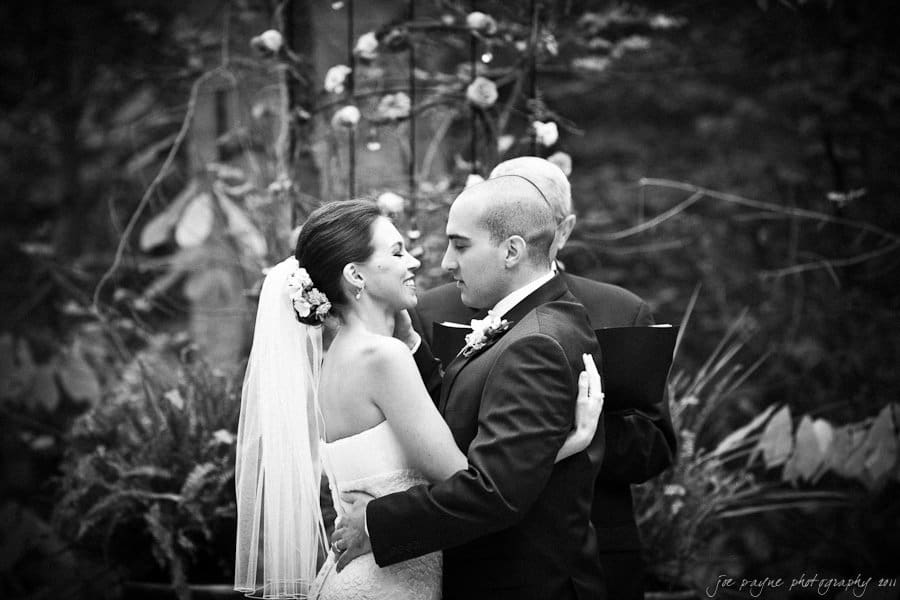 A bride and groom share an intimate moment during their outdoor wedding ceremony. The bride, in a strapless gown with a veil, and the groom, in a suit, gaze into each others eyes, surrounded by flowers and greenery.