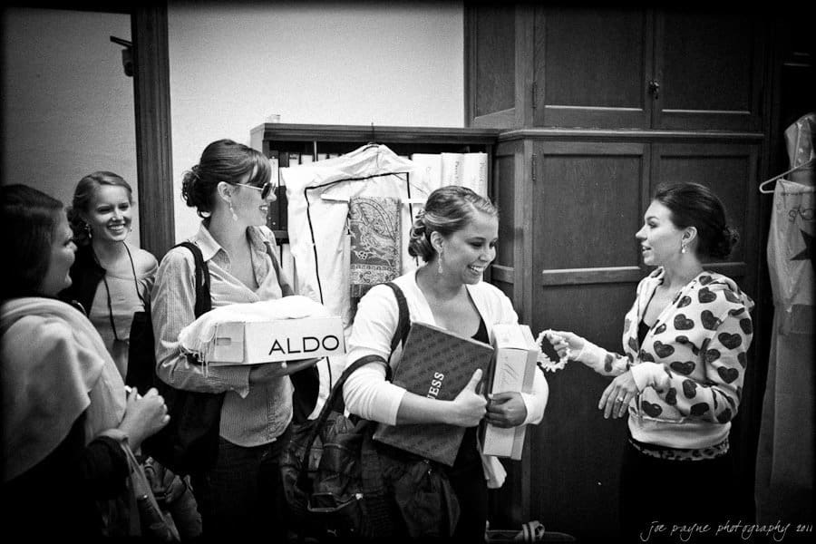 A group of women smiling and talking indoors. One woman holds boxes and a book, another carries a garment on hangers covered in plastic. Shelves and a wardrobe are in the background. Black and white image.