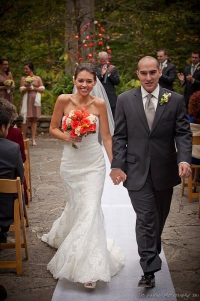 A bride and groom walk down an outdoor aisle lined with guests sitting in chairs. The bride holds a bouquet of orange and white flowers and wears a white lace gown, while the groom is in a dark suit with a boutonniere. Bridesmaids stand in the background.