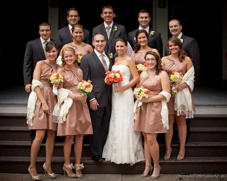 A wedding party poses on steps. The bride and groom stand in the center. Bridesmaids in tan dresses hold orange flower bouquets, and groomsmen in dark suits stand around them. Everyone is smiling at the camera.