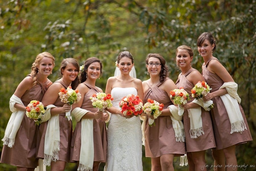 A bride in a white dress stands outdoors with six bridesmaids wearing brown dresses. They are all holding bouquets of orange and white flowers, with greenery in the background.