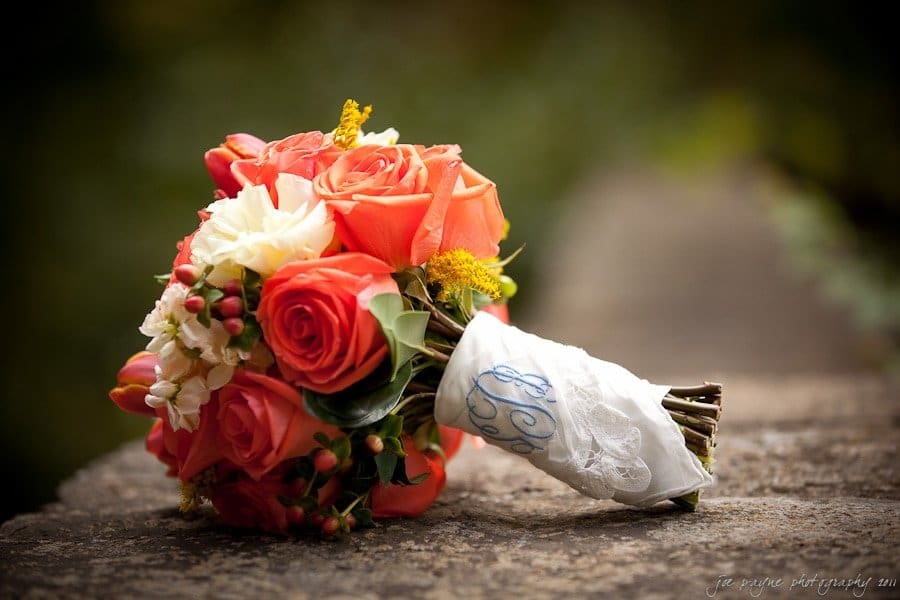 A vibrant bridal bouquet with pink, red, and white roses, along with small yellow flowers and greenery, is wrapped in white lace and placed on a stone surface.