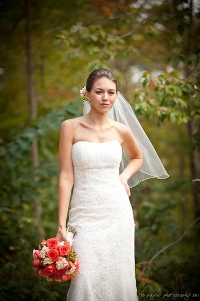A woman in a white strapless wedding dress and veil stands outdoors, holding a bouquet of red and pink flowers. She is surrounded by lush greenery, creating a serene and natural backdrop.
