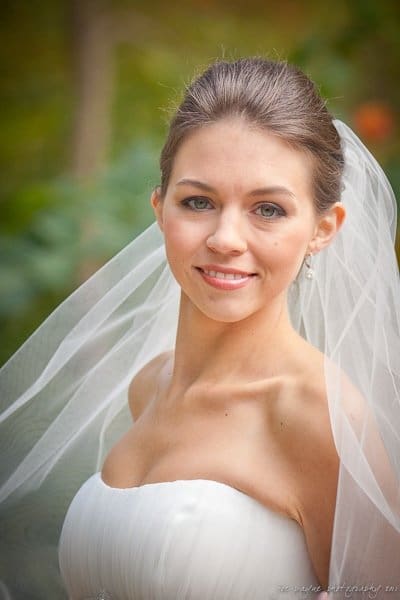 A bride with a white veil smiling at the camera. She is wearing a strapless white dress with her hair neatly styled. The background is blurred greenery.