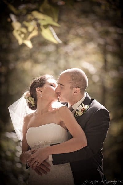 A bride and groom share a kiss in an outdoor setting. The bride is wearing a white dress and veil, and the groom is in a dark suit with a boutonniere. They are surrounded by soft, natural light and blurred greenery in the background.