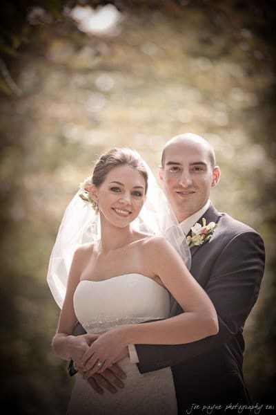 A bride and groom stand outdoors, smiling. The bride wears a strapless white gown and veil, while the groom wears a dark suit with a boutonnière. They are embracing, with soft lighting and blurred greenery in the background.