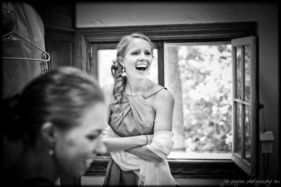 A woman in a sleeveless dress is laughing and standing near a window. Another person is partially visible in the foreground. The setting appears casual and cheerful, with natural light coming through the window.