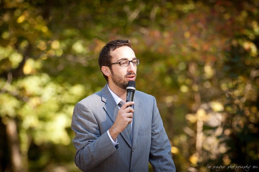 A man in a gray suit and glasses is speaking into a microphone outdoors. He is standing against a backdrop of blurred green foliage.
