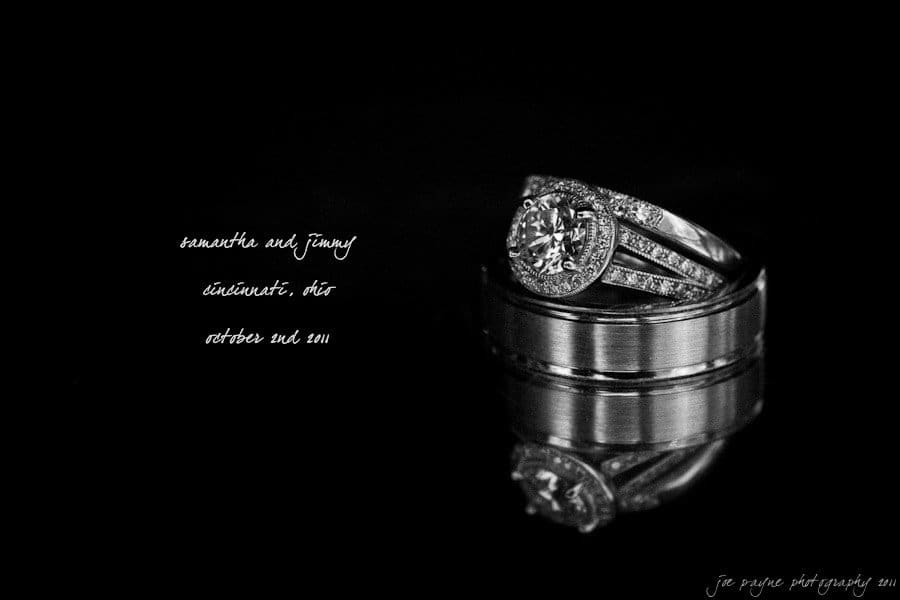 A close-up of two wedding rings against a black background. One ring is adorned with a large diamond and smaller diamonds on the band, while the other is a plain silver band. Handwritten text reads: Kenneth and Ginny, Cincinnati, Ohio, October 2nd, 2011.
