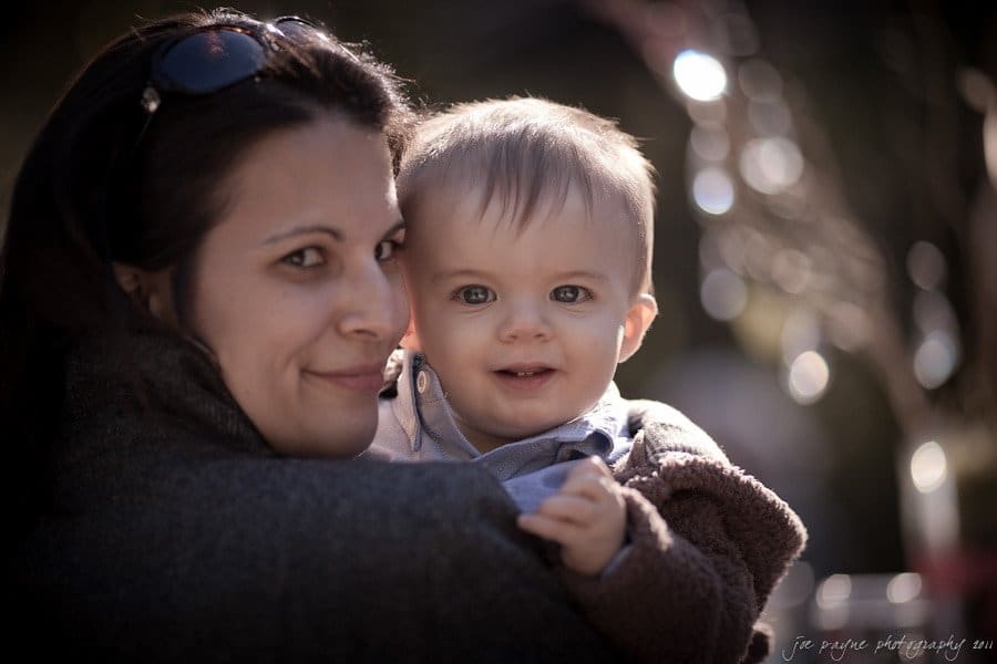 A woman holding a baby in her arms, both smiling at the camera. The background is softly blurred, highlighting their close and cheerful bond. The sunlight creates a gentle halo effect around them.