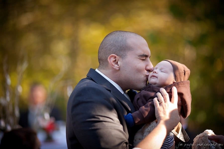 A man in a suit gently kisses a baby wearing a brown outfit. The scene is set outdoors with blurred greenery in the background, suggesting a warm, intimate moment. The lighting is soft and natural.