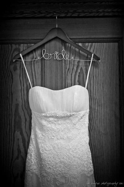 A black and white image of a strapless wedding dress hanging on a wooden hanger. The hanger is labeled with the word bride in wire script. The dress features lace detailing on the bodice and skirt.