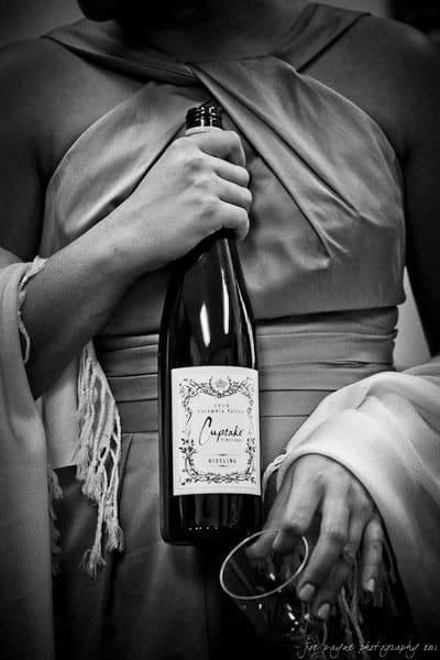 A person in a formal dress holds a bottle of Cupcake wine and an empty glass. The grayscale image highlights the elegant, draped fabric of the dress. The persons face is not visible.