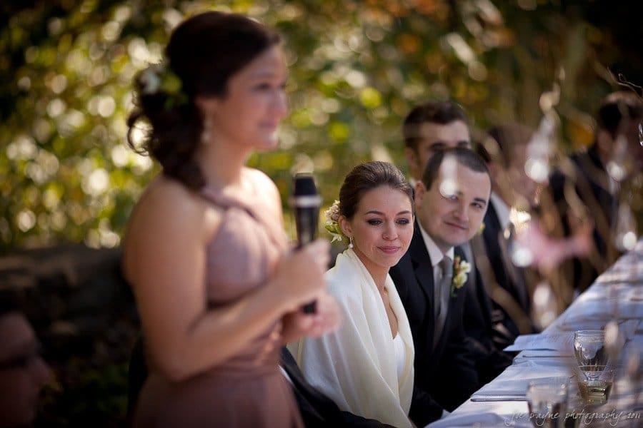 A bridesmaid in a pink dress speaks into a microphone at an outdoor wedding reception. The bride, wearing a white shawl, and a groomsman sit at a long table, listening attentively. The background is filled with greenery.