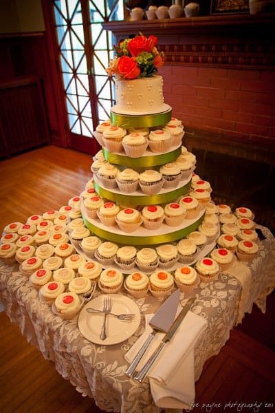 A tiered display of cupcakes with a small cake on top, decorated with orange and green flowers. The cupcakes are arranged on a lace-covered table and are frosted in ivory with colorful centers. Silverware and napkins are set nearby.