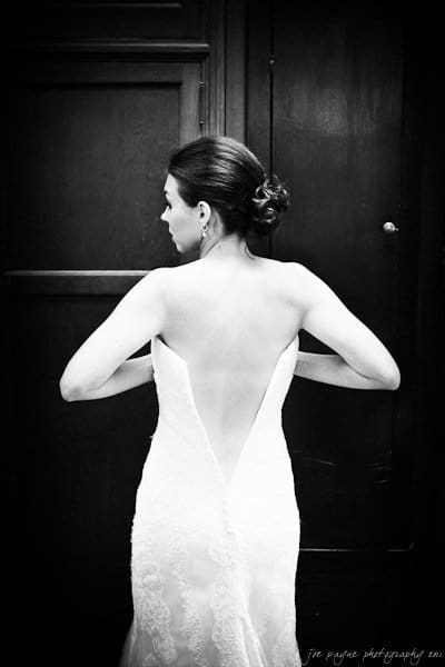 A black and white photo of a woman in an elegant wedding dress with a deep V back. Her hair is styled in a neat bun, and she stands with her hands on her hips against a dark paneled wall.