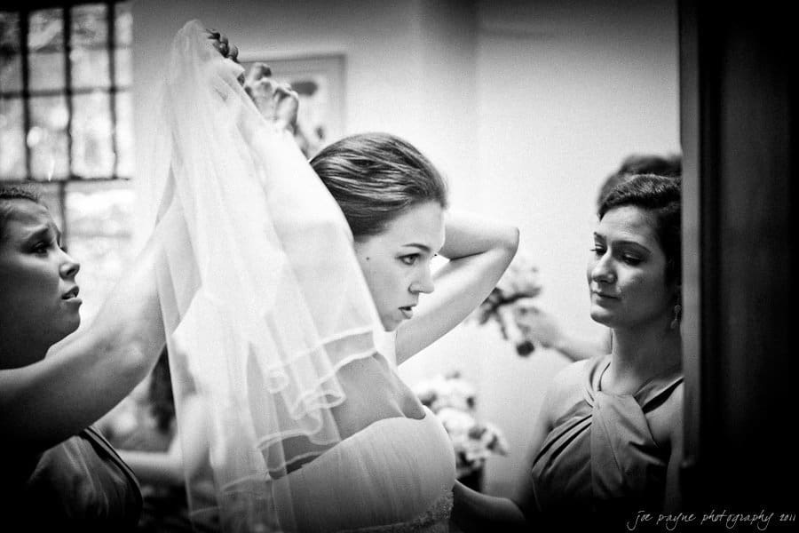 A black-and-white photo of a bride being helped by two bridesmaids as she adjusts her veil. The bride appears focused, while the bridesmaids assist with care. They are in a softly lit room with a window in the background.