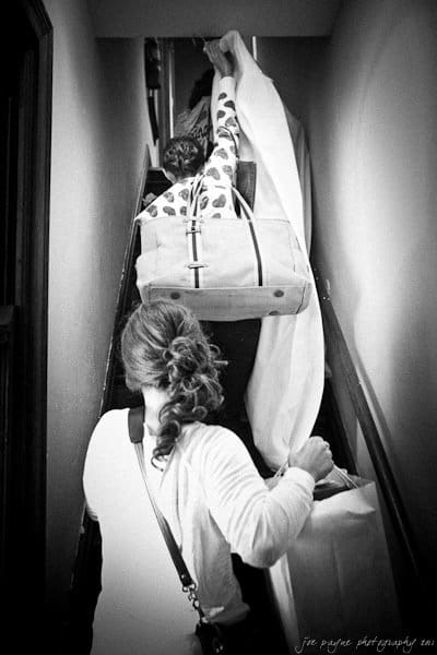 Two people carrying large bags and holding a draped fabric climb a narrow staircase. The photo is in black and white, emphasizing the confined space and the effort involved in moving the objects.