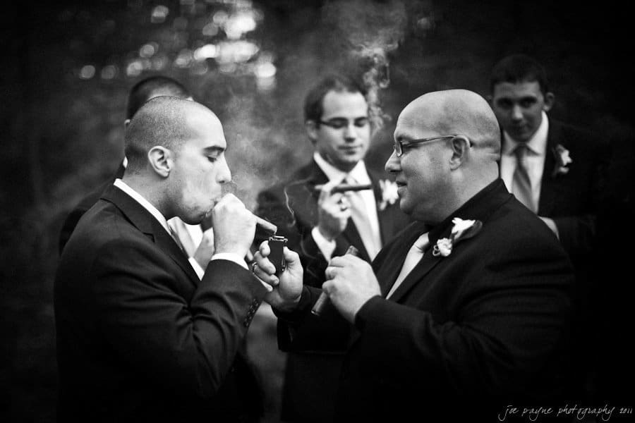A group of men in suits stand together outdoors, all appearing to be groomsmen. One man lights a cigar for another. Smoke wafts through the air, and they look relaxed and celebratory.