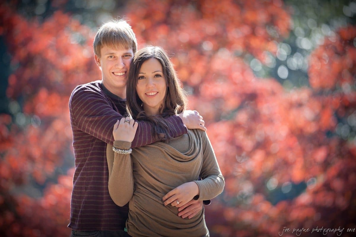 A couple stands together outdoors, smiling, with the man hugging the woman from behind. They are surrounded by vibrant autumn foliage with red leaves in the background.