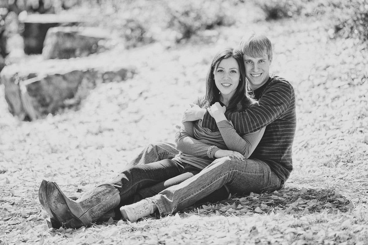 A couple sitting on a leafy ground, smiling at the camera. They are wearing casual clothes and embracing each other. The background is filled with blurred trees and rocks, creating a peaceful outdoor setting.