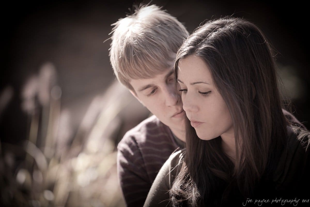A couple sits outdoors, with the man gently embracing the woman from behind. She gazes downward with a thoughtful expression. The sunlight casts a soft, warm glow on their faces, creating a serene and intimate atmosphere.