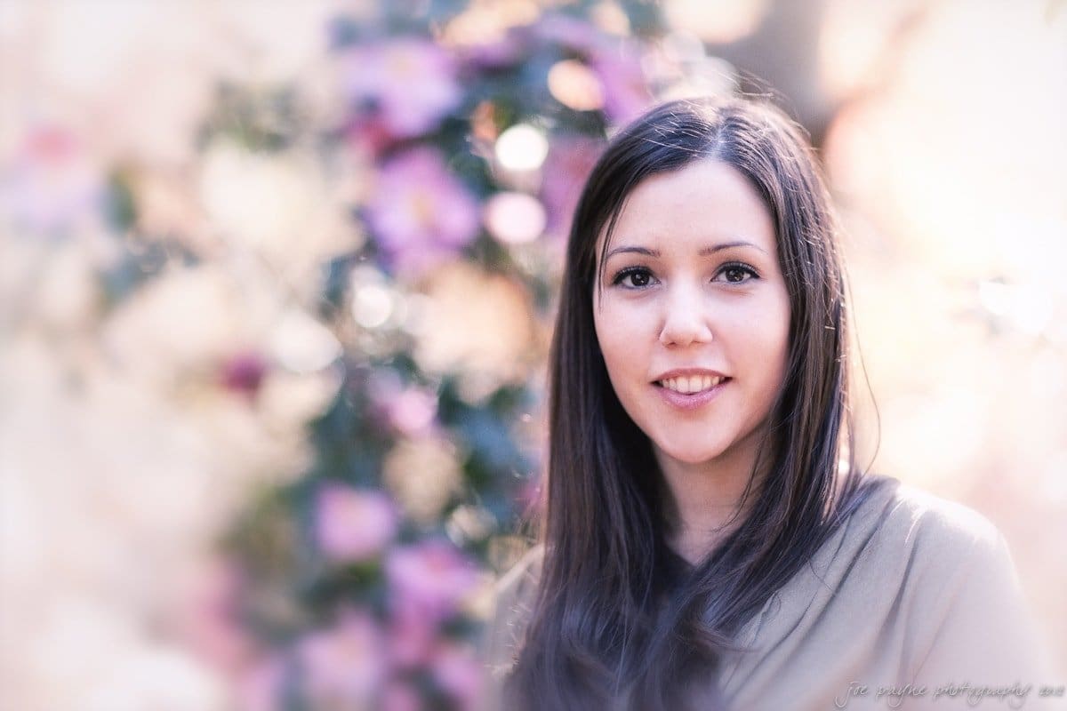 A woman with long brown hair smiles softly while standing outdoors. The background is filled with soft-focus pink and purple flowers, creating a dreamy atmosphere.