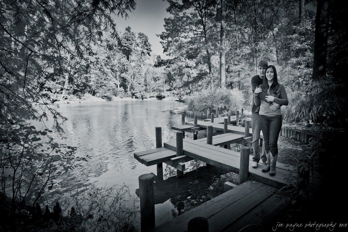 A black and white image shows a couple walking on a wooden boardwalk along a tranquil lake surrounded by dense trees. The water is calm, and the couple appears to be engaged in a conversation while enjoying the natural scenery.