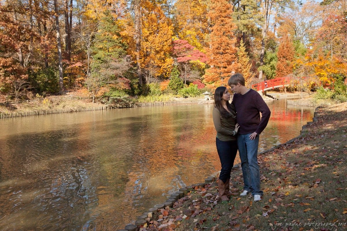 A couple stands by a serene lake surrounded by autumn trees with vibrant orange, red, and yellow leaves. They are embracing and smiling, while a red bridge is visible in the background. The ground is covered with fallen leaves.