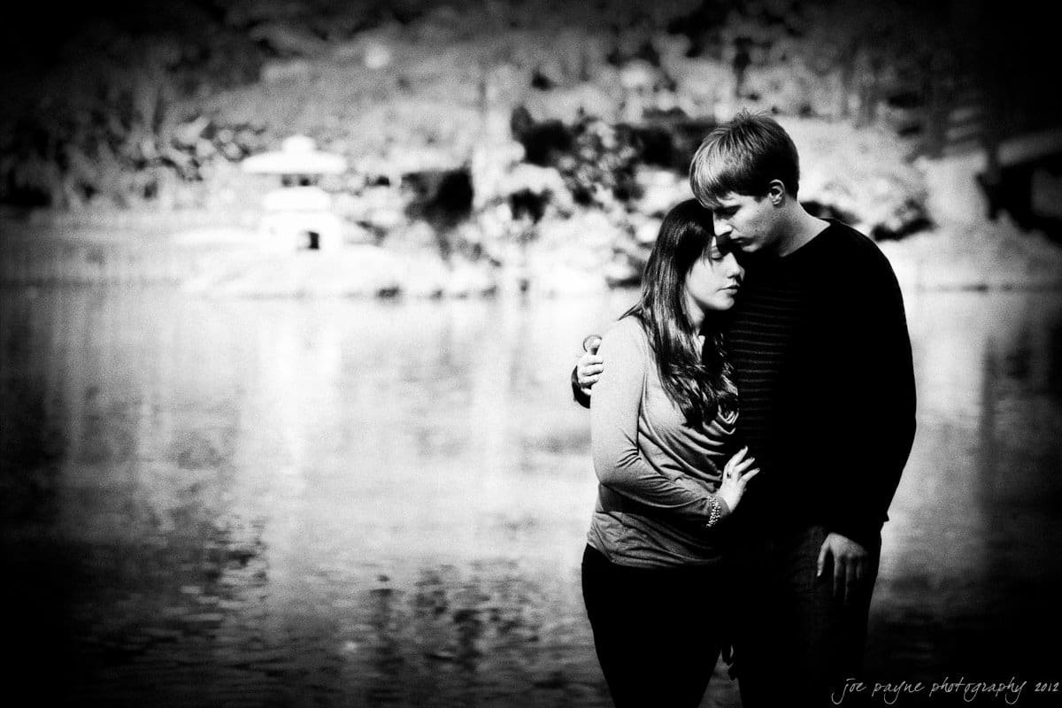 A couple stands by a tranquil lake, embracing tenderly. The woman leans her head on the mans shoulder, eyes closed. The scene is captured in black and white, with a soft focus on the serene water and blurred background scenery.