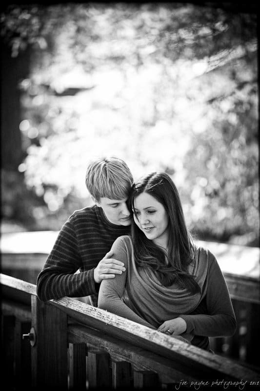 A black and white photo of a couple on a wooden bridge. The man stands behind the woman, gently resting his arm on her shoulder. They both look content and are surrounded by a blurred natural background.