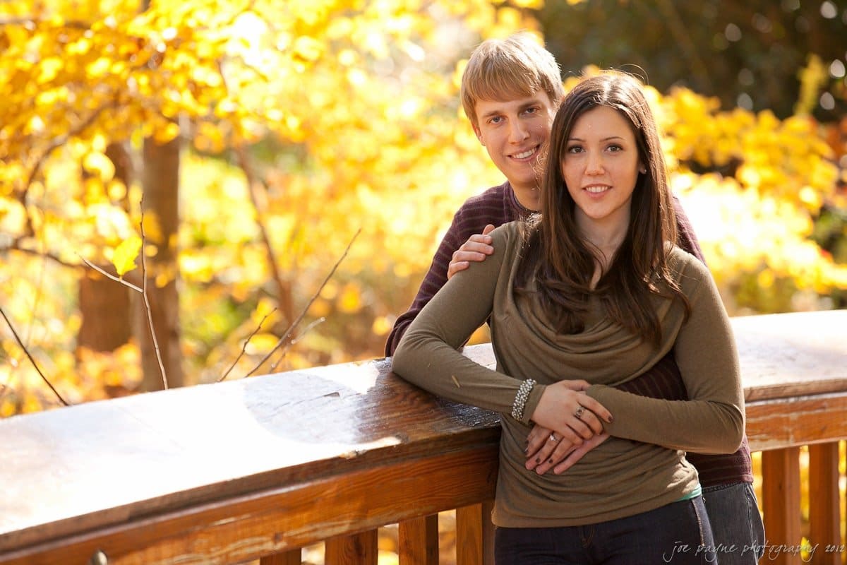 A couple stands on a wooden bridge surrounded by vibrant autumn foliage. The woman, wearing a brown top, leans on the railing with a smile, while the man stands slightly behind her, smiling. Golden leaves create a warm background.