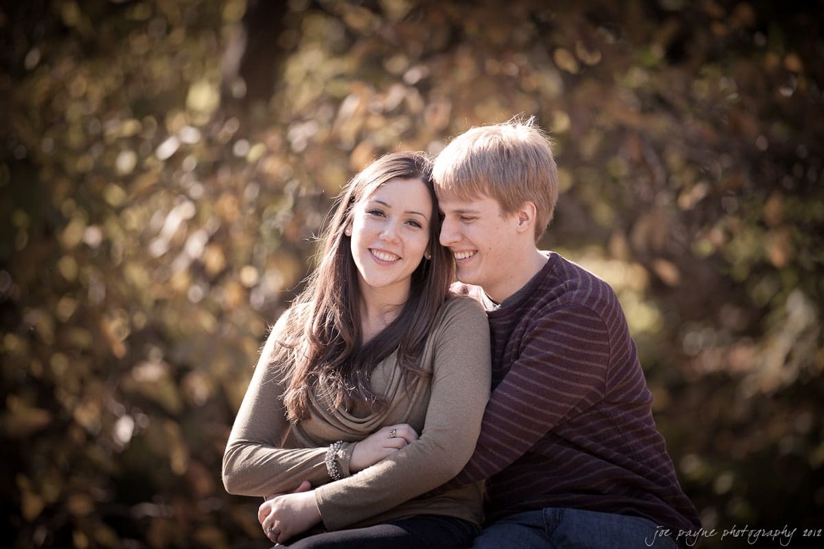 A young couple sits together outdoors with autumn foliage in the background. The woman smiles gently, while the man leans in, appearing happy and content. They are dressed casually in long-sleeve shirts.