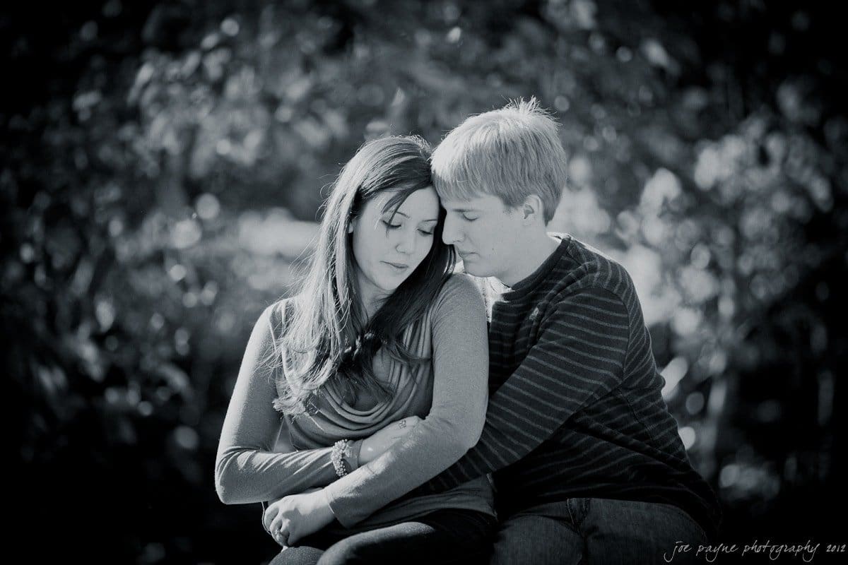 A black and white photo of a couple sitting close together, surrounded by blurred foliage. The woman gazes down, and the man leans in with his eyes closed, creating an intimate and peaceful scene.