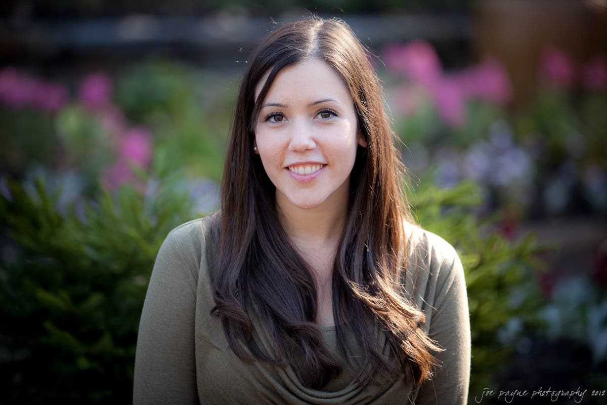 A woman with long brown hair smiles at the camera, standing in front of a blurred garden background with pink and green plants. She is wearing a brown top.