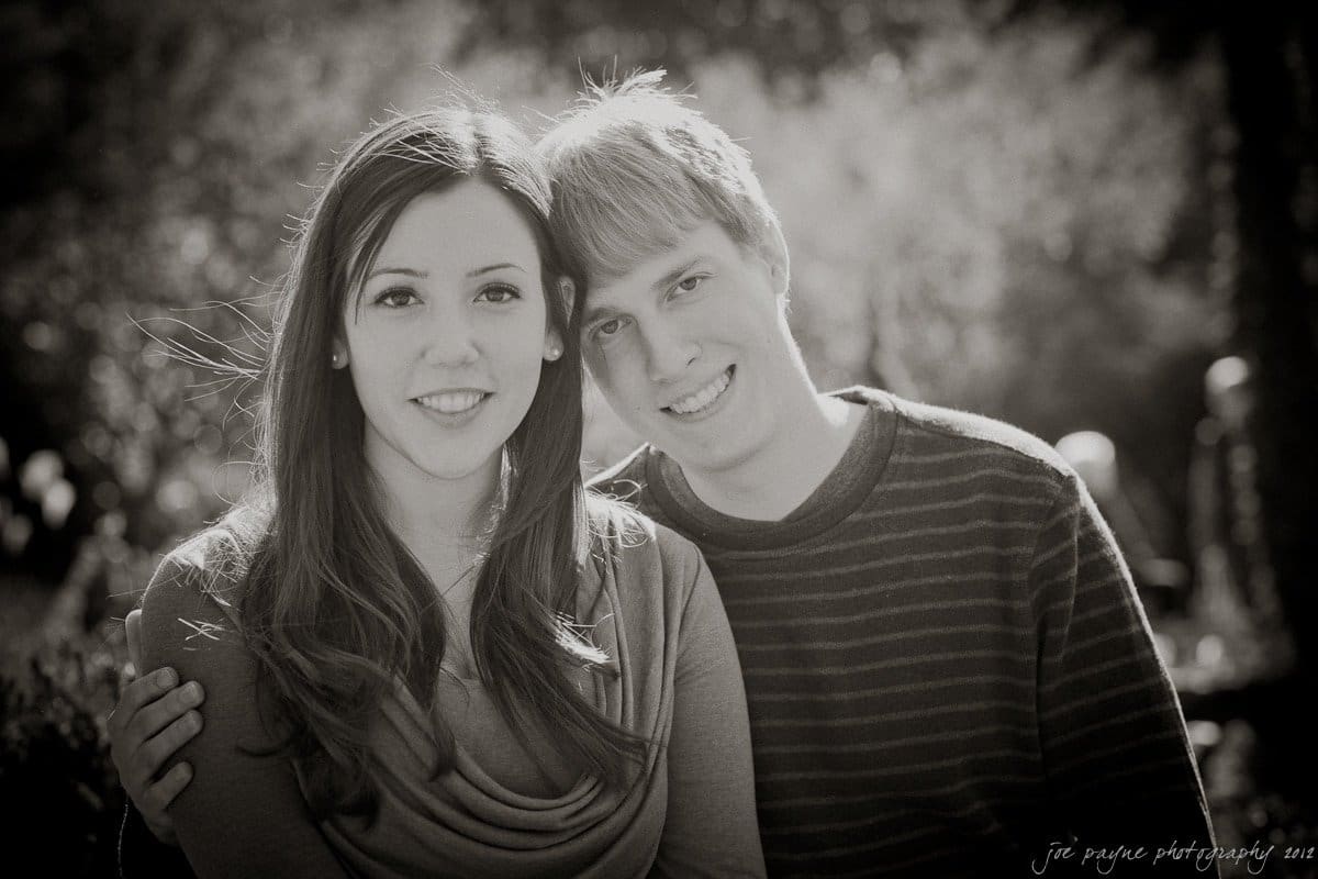 A couple sits closely together outdoors, smiling at the camera. The woman has long hair and wears a draped top, while the man has short hair and wears a striped sweater. The background features sunlight filtering through trees, creating a serene setting.