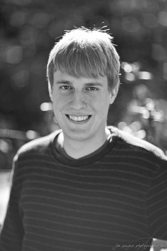 A black-and-white portrait of a smiling person with short, light-colored hair wearing a striped sweater. The background is blurred, creating a soft bokeh effect.
