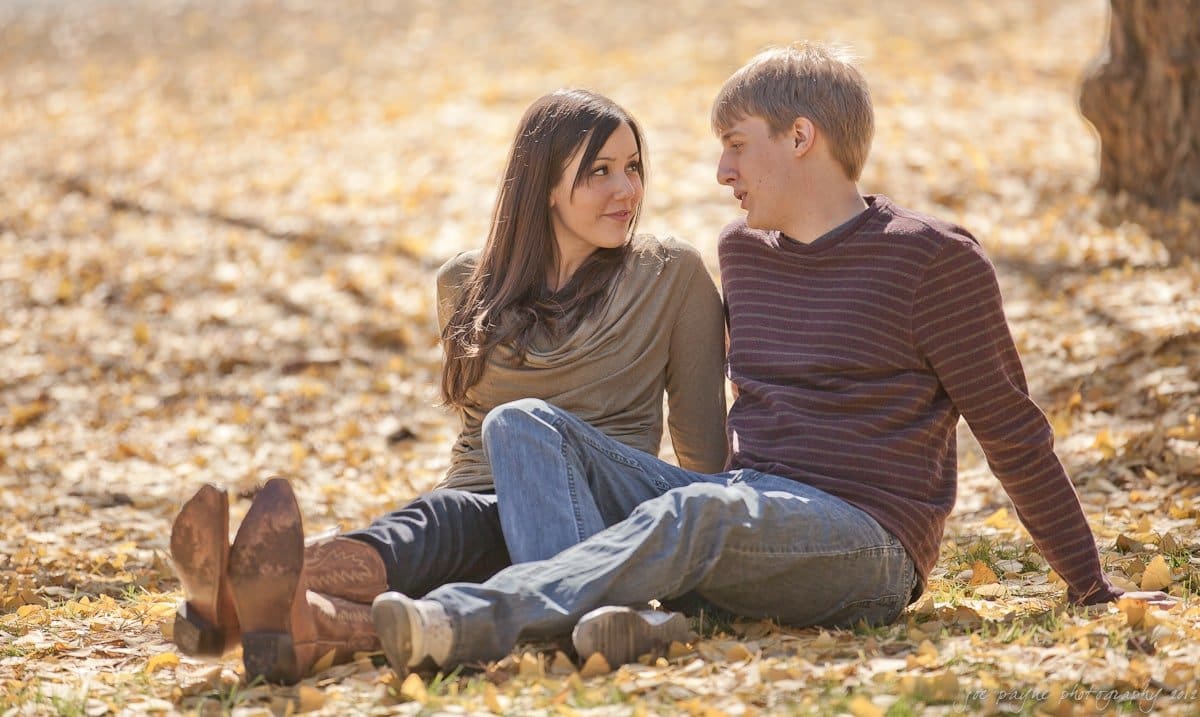 A man and woman sit on a blanket in a park covered with fallen leaves, looking at each other. The woman wears brown boots and a green sweater, while the man wears a striped sweater. Sunlight filters through the trees, creating a warm, autumn atmosphere.