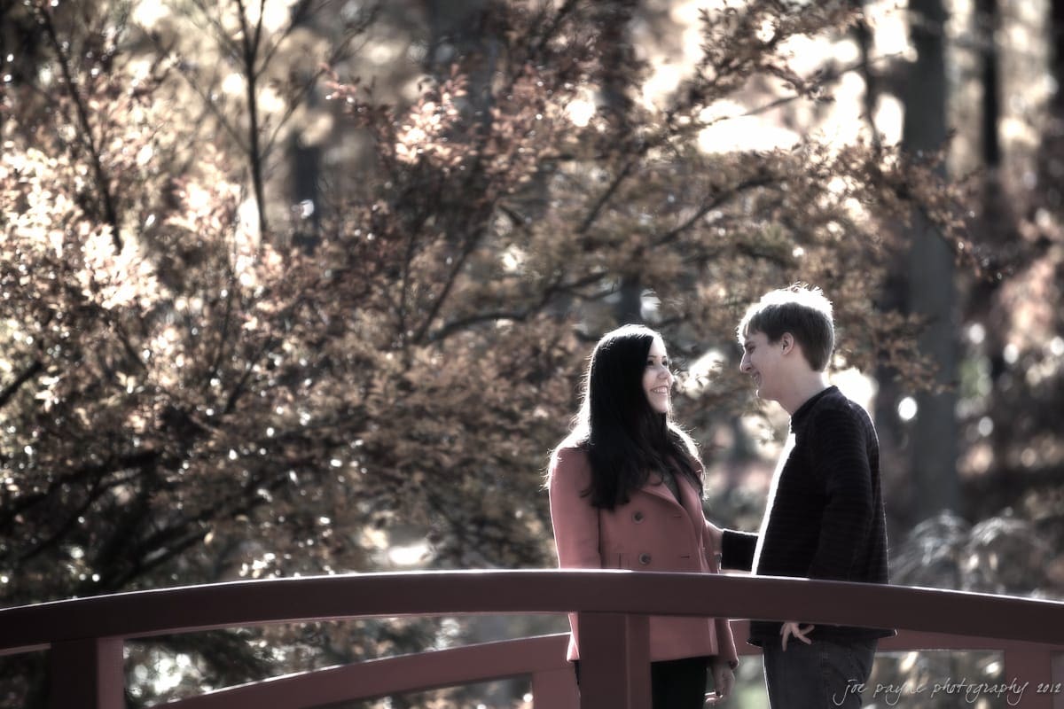 A woman and a man stand on a bridge in a park, smiling at each other. The background features autumnal trees with golden leaves, creating a warm, serene atmosphere.