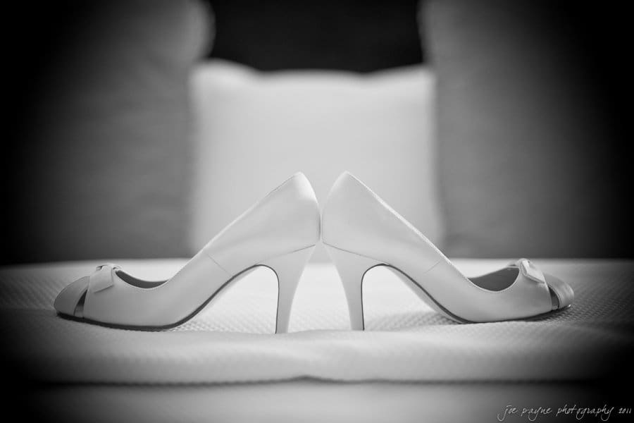Black and white image of a pair of high-heeled shoes facing opposite directions, placed on a soft textured surface with blurred pillows in the background.